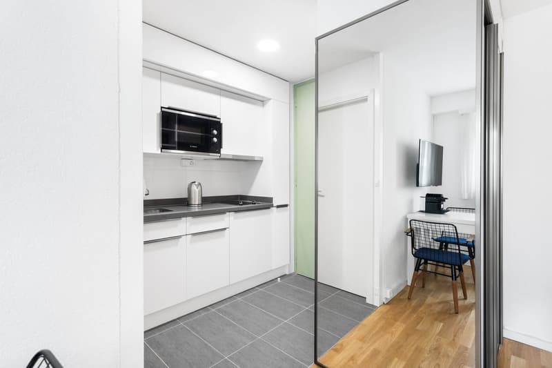 Modern, open-concept studio apartment with a compact kitchen featuring white cabinets, a built-in microwave, and a dishwasher. The apartment has a tiled floor in the kitchen area and hardwood floors in the living space. There is a small table and chair se