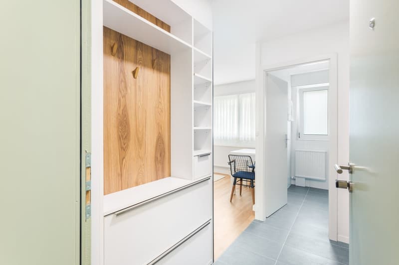 The image shows a modern and minimalist hallway with a white built-in storage unit featuring wooden accents. The hallway leads to a room with a chair visible through the open doorway. The floor is covered with gray tiles, and the walls are painted white,