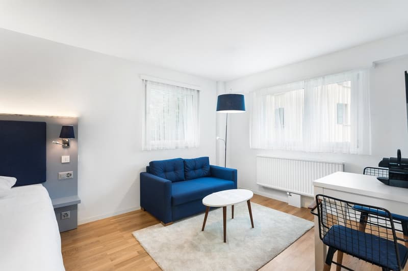 Spacious living room with white walls, large windows, blue sofa, coffee table, and floor lamp. Hardwood flooring throughout the room.