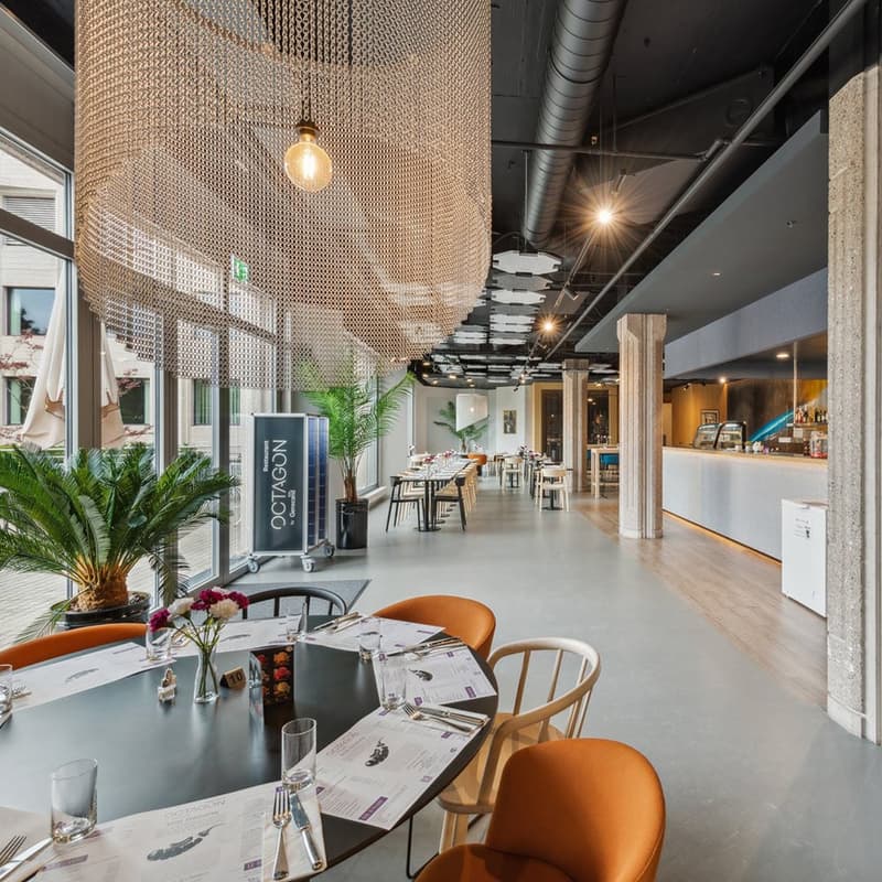 Contemporary style restaurant with glass walls, wooden tables, and chairs, chandeliers, tiled floor, reception counter, tables with flower arrangements, and menus.