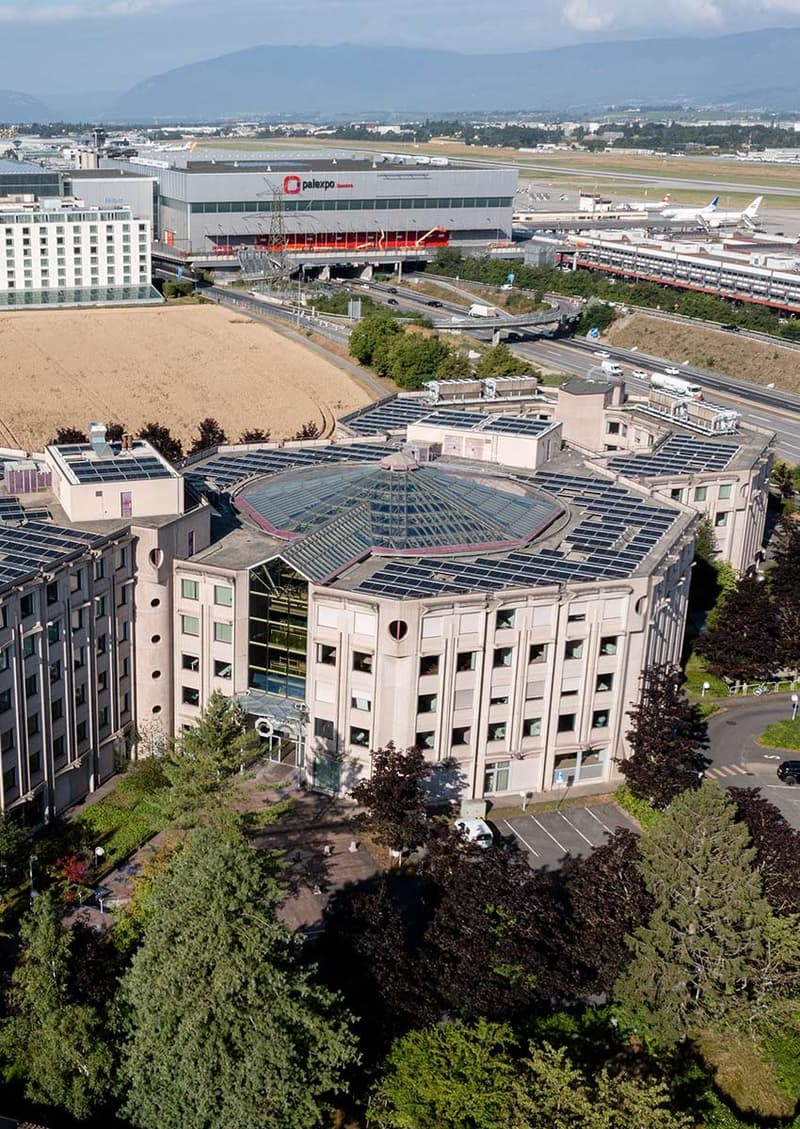 Modern, multi-story building, solar panels on rooftop, surrounded by trees and plants, nearby parking lot