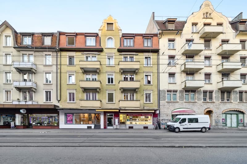 Three multi-story apartment buildings on a street corner, one with a sign reading 'Stargold Uhren - Bijouterie', and one white van parked in front of the middle building.