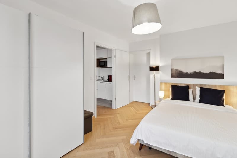 white walls, wooden floors, bed with white bedding and black pillows, open closet, a small bedroom, a small kitchen with a microwave, sink, and refrigerator