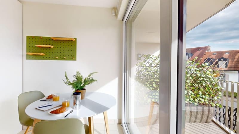 A small dining area with a table, chairs, and a balcony with plants. There is a mounted green pegboard on the wall.