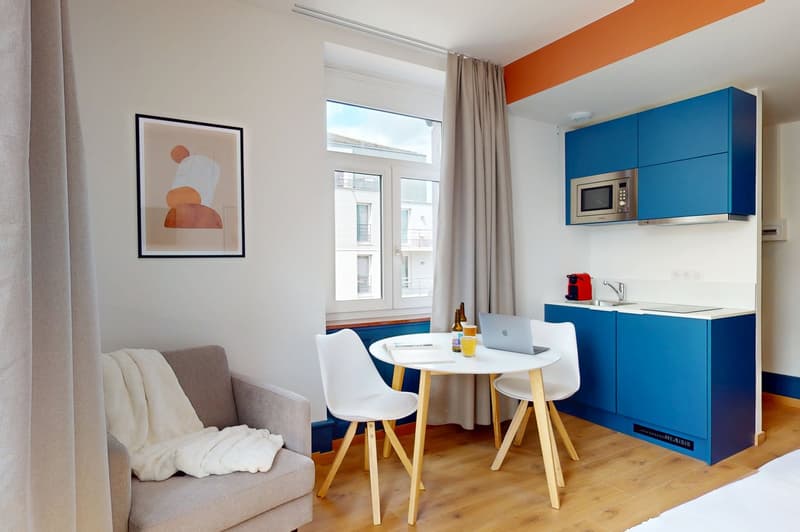 Cozy room with white walls, wooden floor, small table, white chairs, gray couch, framed picture, blue kitchen cabinets, microwave, sink, faucet, microwave, wine glasses.
