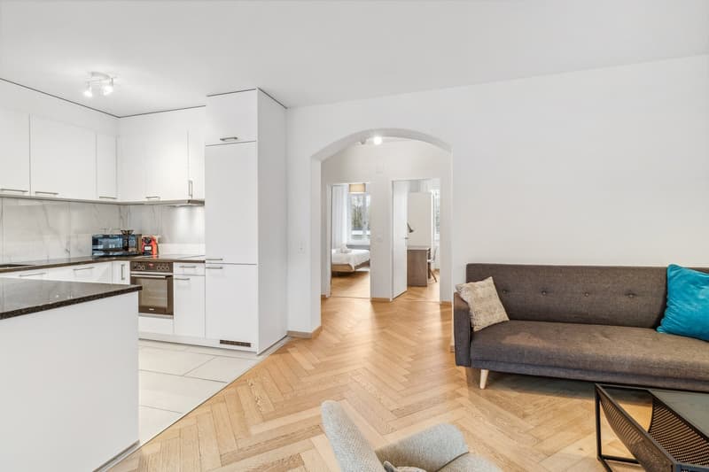 living room with modern furniture, open-plan kitchen, parquet flooring, arched entrance, beige sofa, white pillows, armchair, glass coffee table, white cabinets, countertop, sink, oven