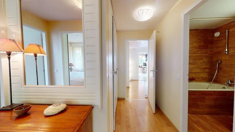 corridor with a large mirror, hardwood floors, large windows, wooden desk with a lamp, hallway leading to a bathroom with a bathtub