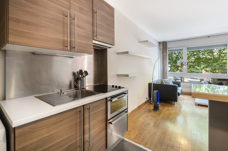 modern kitchen with wooden cabinets, stainless steel appliances, white countertops, black stove, sink, oven, and a small couch in the living area