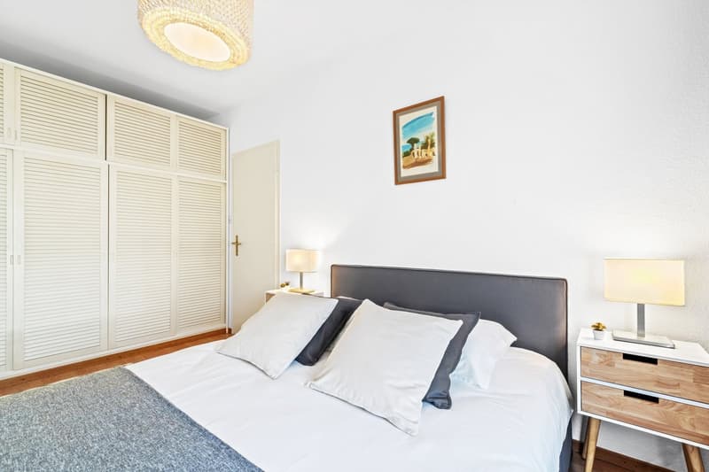 Spacious bedroom with white walls, hardwood floors, and a large wardrobe. The room features a king-size bed with white bedding and gray accents, a bedside table with a lamp, and a framed artwork on the wall. There is also a balcony or terrace visible thro