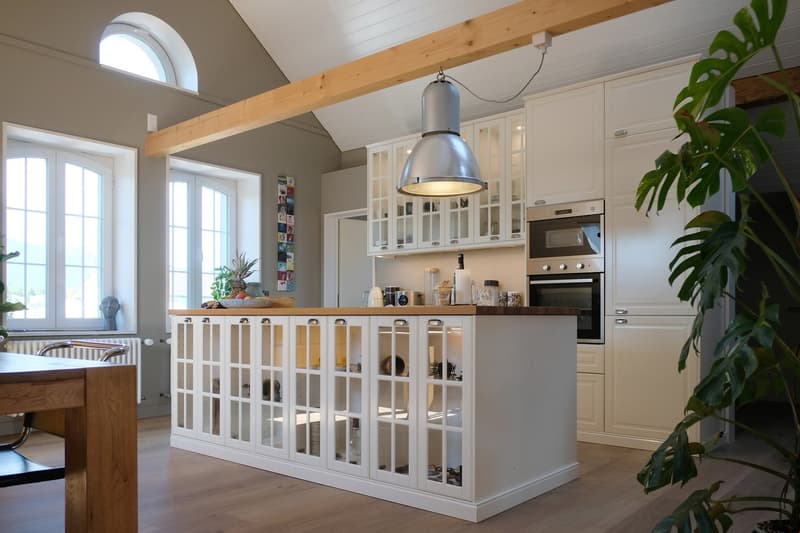 Modern kitchen with open concept layout, white cabinetry, wooden countertop, white island with glass doors, modern lighting fixture, white appliances, wooden dining table, wooden ceiling beams, windows with white frames.