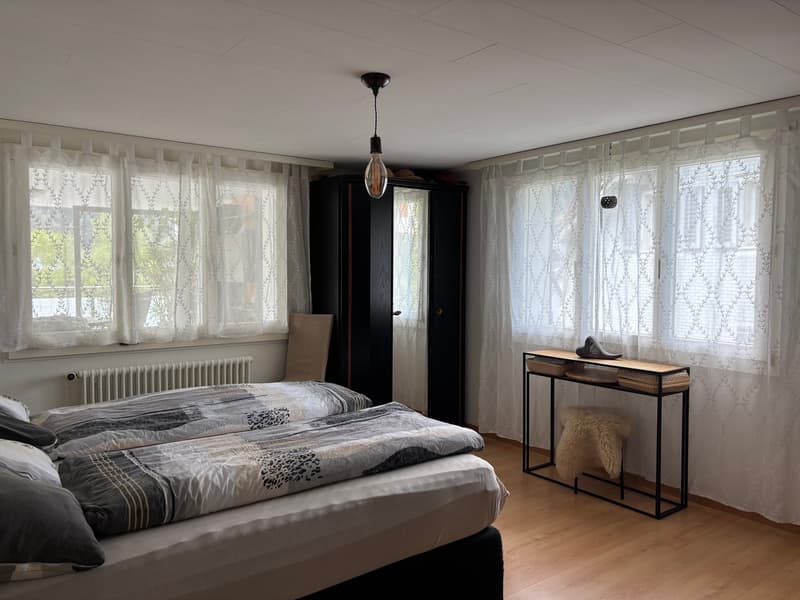 Two beds, white curtains, wooden floor, wardrobe, radiator, table with fur stool