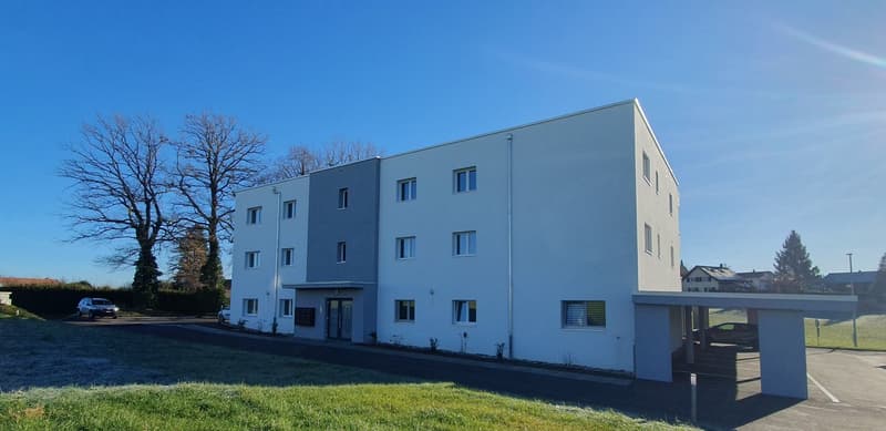 A three-story residential building with white exterior walls, several windows, a covered entrance, and a large driveway in front of it. A vehicle is parked under the covered entrance. There are cars parked in the parking lot on the right side of the build