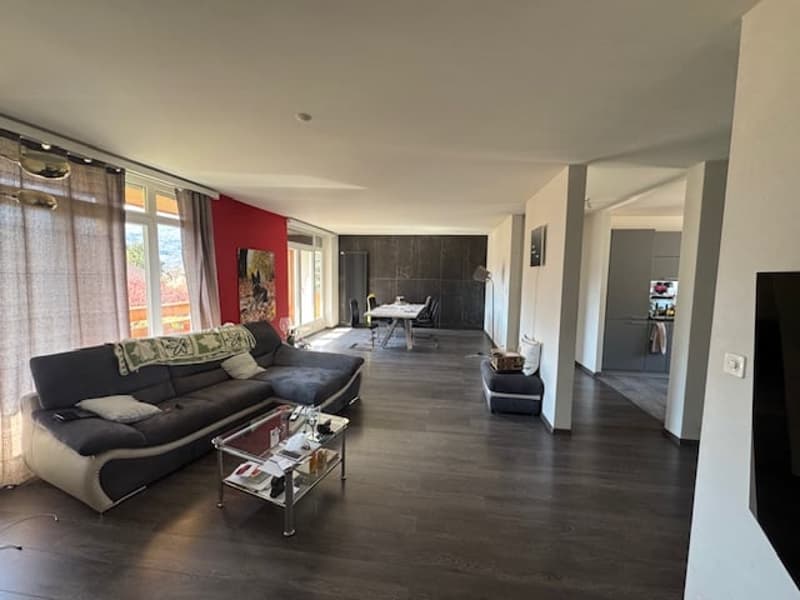 modern living room with gray sofa, glass coffee table, wooden floor, large windows, dining table, and kitchen