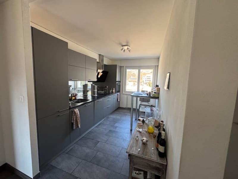 modern kitchen with gray cabinets, tiled floor, stainless steel appliances, a window with a view, a wooden table with bottles and a yellow object