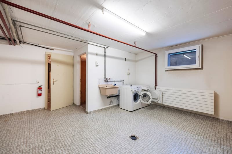 This image shows a laundry room with a washing machine and dryer, a sink, and tiled flooring. The room has white walls and exposed pipes and wiring on the ceiling. There is a window and a door leading to another room.