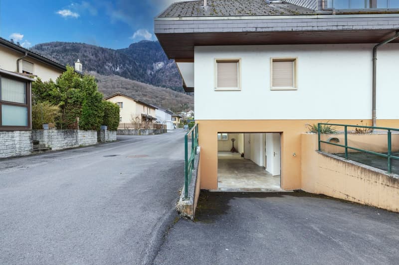 2-story residential building with a garage, surrounded by trees and mountains in the background. The building has a white exterior with green railings and a slanted roof.