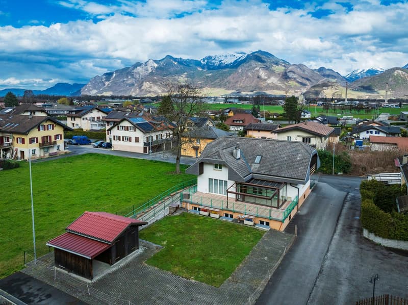 The image shows a scenic view of a small town nestled in a mountainous landscape. The town features colorful houses with red tile roofs, surrounded by green fields and trees. In the background, there are snow-capped mountains under a blue sky with fluffy