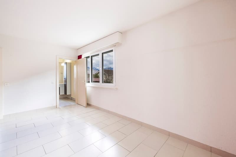 This image shows an empty room with white walls and a large window overlooking what appears to be a mountainous landscape outside. The room has a tiled floor and a doorway leading to another area. The room appears to be clean and well-lit, with a minimali