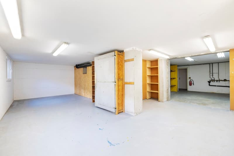 Large open space with concrete floor, white walls, and various storage units and shelves. There appears to be a garage door or entrance on the left side of the image.