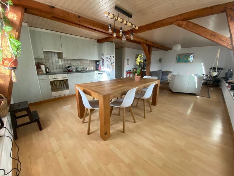 Dining room with wooden table, white chairs, wooden beams, kitchen with appliances, living room with couch, plants, and decorations