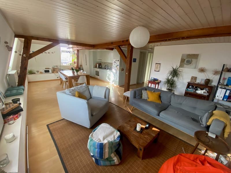 Open living room with wooden floor, wooden beams, white walls, two sofas, wooden coffee table, two armchairs, carpet, dining table, chairs, kitchen, microwave, refrigerator, potted plants, shelves, decorative items