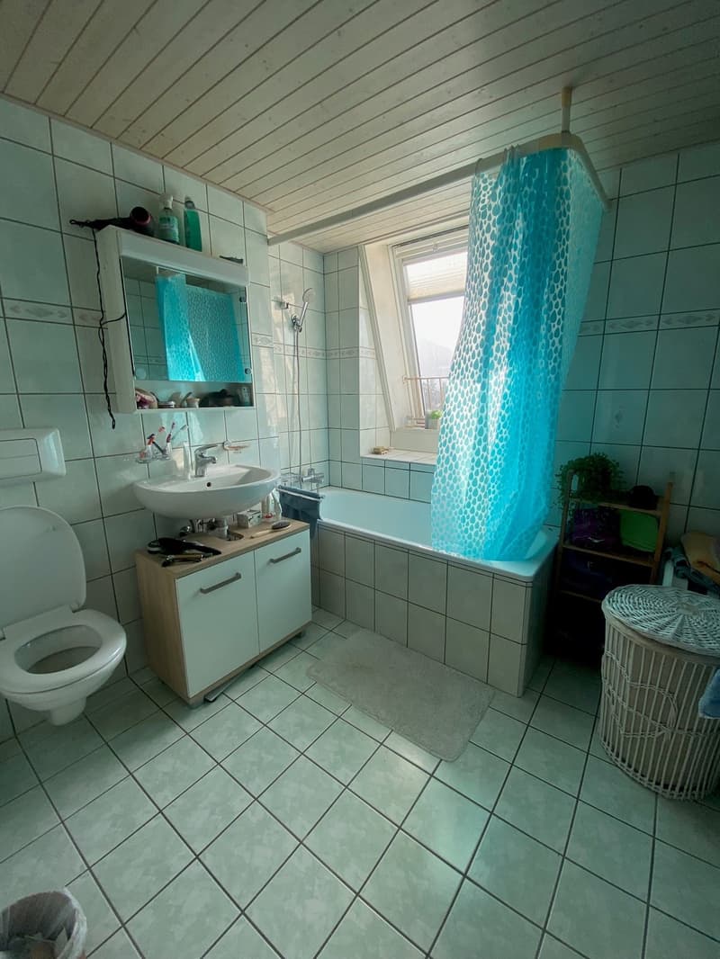 Bathroom with white tiles, white toilet, white sink, bathtub, blue shower curtain, wooden cabinet, mirror, and a window.