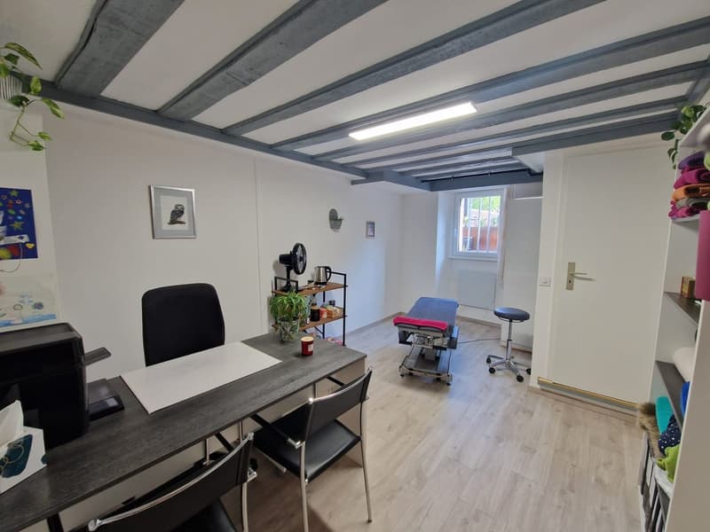 A room with a desk, chairs, a fan, a shelf, a stool, a massage table, and a window.