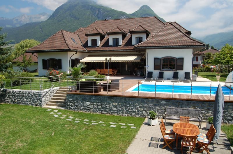 Large two-story house with a tiled roof, surrounded by mountains. The house has a patio with a swimming pool, wooden deck, and outdoor furniture. There is a stone wall around the property and a grassy lawn with a paved walkway. The house appears to be in