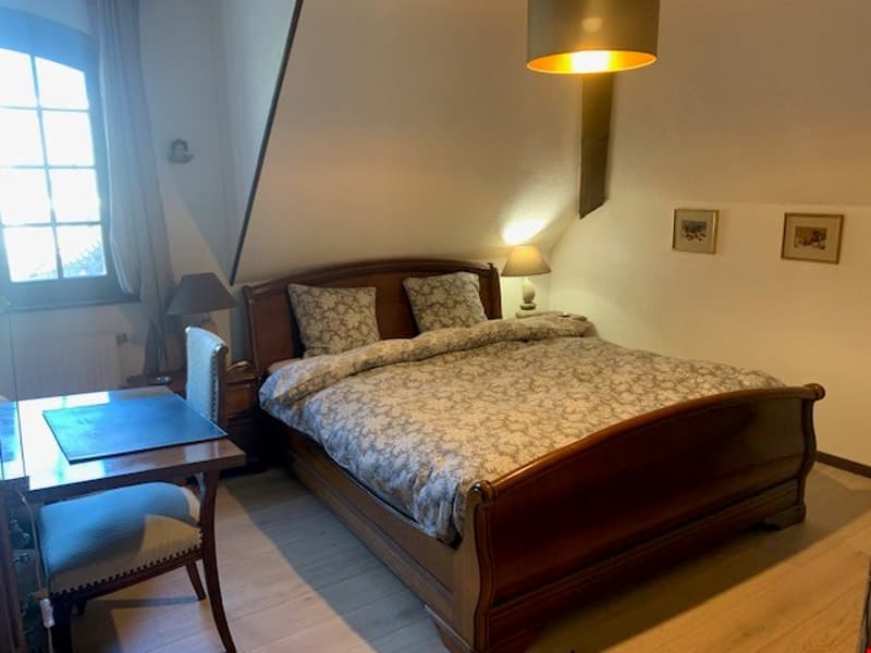 Spacious bedroom with a wooden sleigh bed, floral patterned bedding, a wooden nightstand, and a lamp with a gold shade. The room has a large window and hardwood floors.