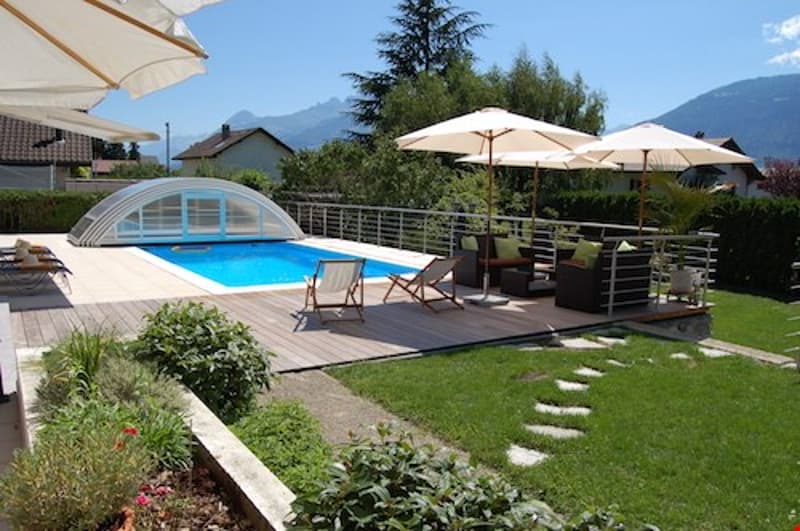 Outdoor swimming pool surrounded by patio furniture and umbrellas, with a grassy lawn and landscaping visible.