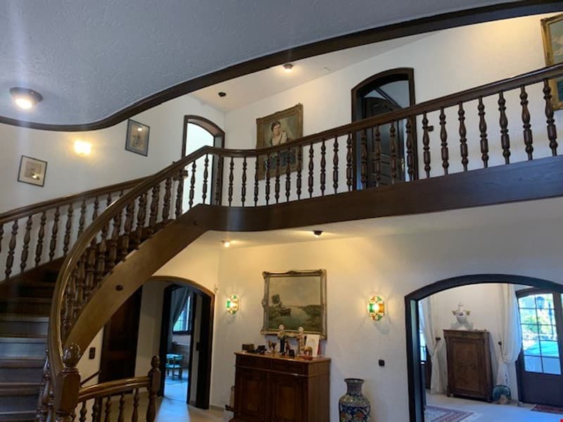 The image shows the interior of a large, two-story house with a grand staircase and balcony. The walls are painted in a light color, and the floors appear to be hardwood. The staircase features ornate wooden railings and spindles. There are several arched