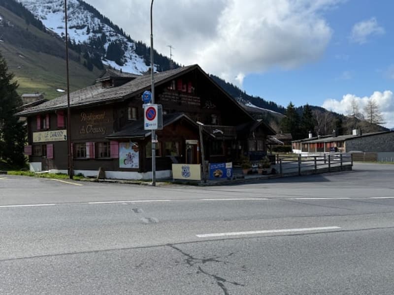 A traditional Swiss chalet-style building with a wooden exterior, located in a mountainous area with snow-capped peaks in the background. The building appears to be a restaurant or cafe, with outdoor seating and a parking area in front.