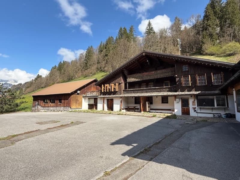 A large wooden chalet-style building with a parking area in front, surrounded by a forested mountainous landscape.