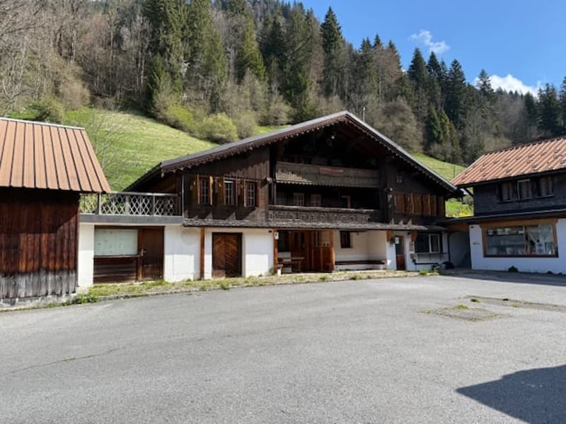 A traditional wooden chalet-style building with a sloped roof, surrounded by a grassy area and trees. The building has a covered porch and several garage doors or parking spaces on the ground floor.