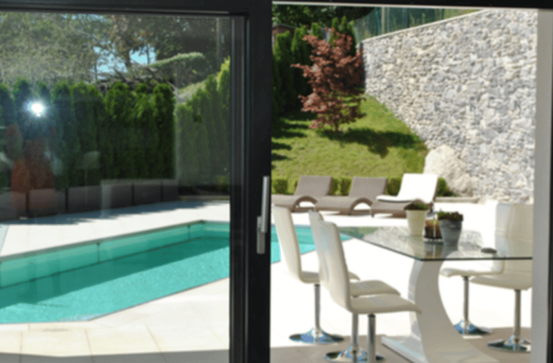 Outdoor patio with a dining table and chairs, overlooking a swimming pool surrounded by a lush garden with trees and shrubs