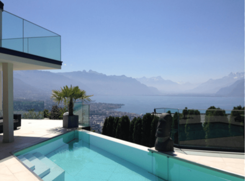 Modern house with a glass balcony, infinity pool, and panoramic view of the mountains and lake