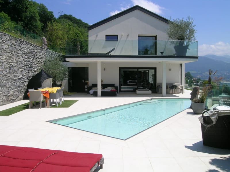 Modern two-story house with a swimming pool, balcony, and glass walls overlooking a scenic landscape. The house has a minimalist design with a white exterior and large windows. There is a patio area with outdoor furniture next to the pool.