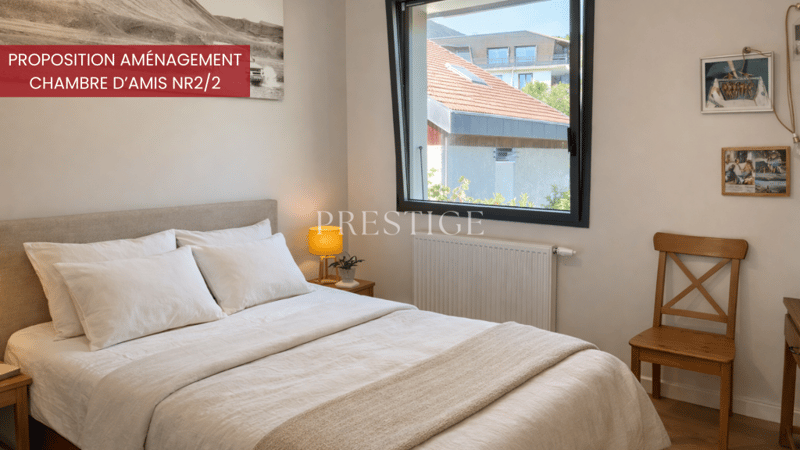 A well-lit bedroom with a window facing the building, bed with white linens, wooden nightstand, lamp, wooden chair, white radiator, framed picture