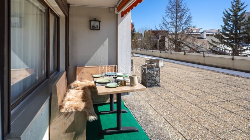 wooden bench, table with plates, fur rug, green carpet, railing, trees, buildings, mountains