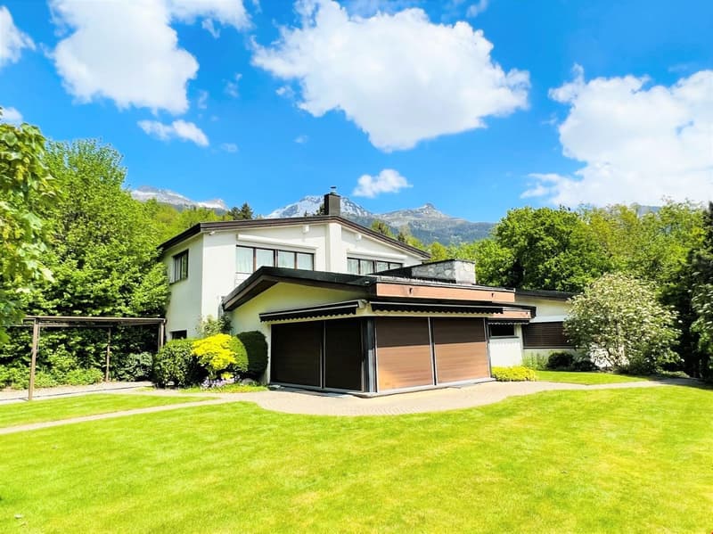 2-story modern house with a garage, surrounded by lush greenery and mountains in the background. The house has a balcony or terrace on the upper level.