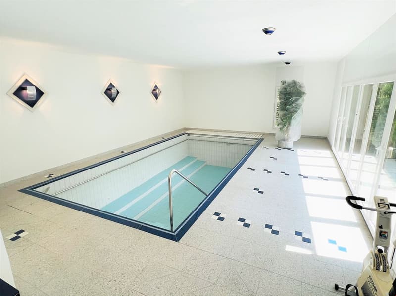 Indoor swimming pool with tiled floor, white walls, and recessed lighting fixtures. The pool has a rectangular shape with a ladder for entry and exit.