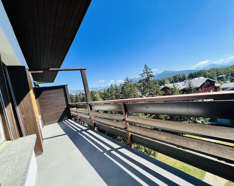 Wooden balcony with railing, overlooking a forested area with mountains in the background under a clear blue sky