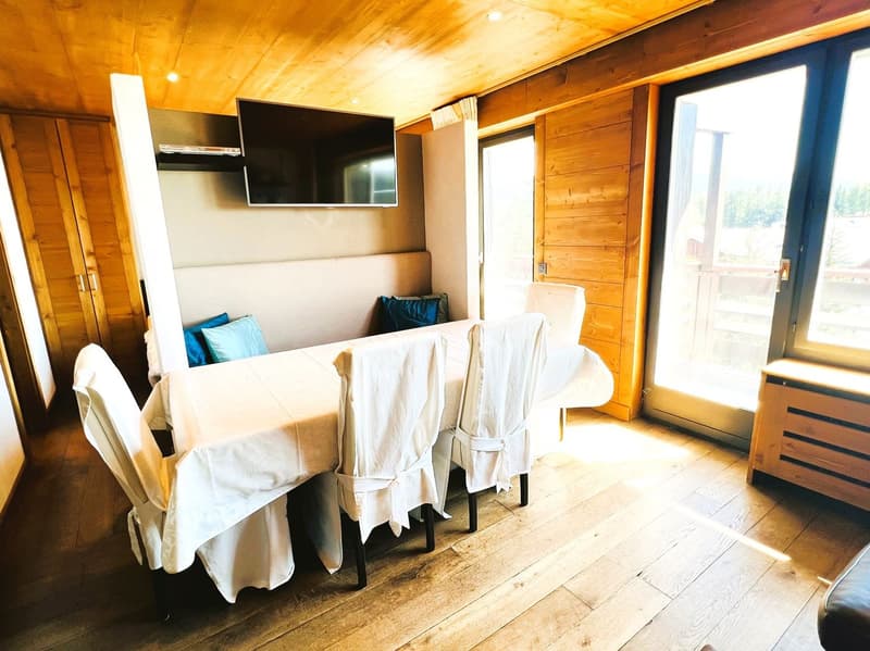 Cozy wooden cabin interior with a dining area, large windows, and a flat-screen TV mounted on the wall. The room has a warm, rustic feel with wooden walls and floors.