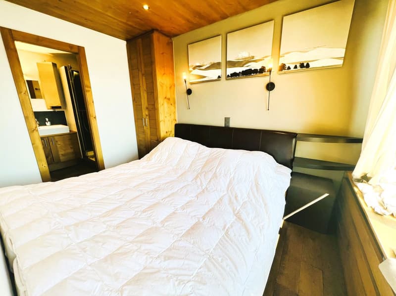 Cozy bedroom with wooden ceiling, white bedding, black leather headboard, and wall art above the bed. There appears to be a fireplace or stove in the room as well.