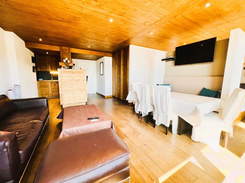 Open-plan studio apartment with wooden ceiling, hardwood floors, leather sofa, dining table, kitchenette, and mounted TV