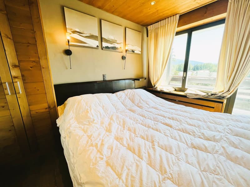 Cozy bedroom with wooden walls and ceiling, large window with a scenic view, king-size bed with white bedding, wall-mounted artwork, and curtains.