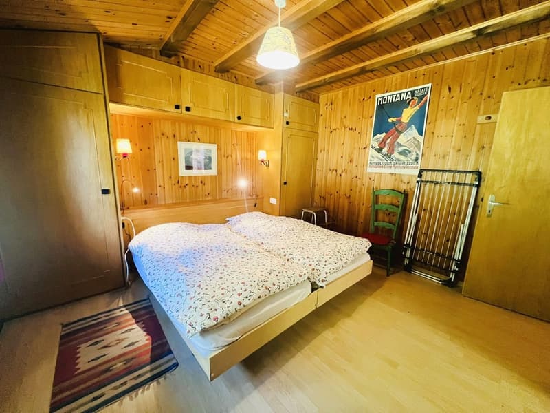 Double bed, wooden wall, bedspread with flowers, wooden chair, open wardrobe, lamp