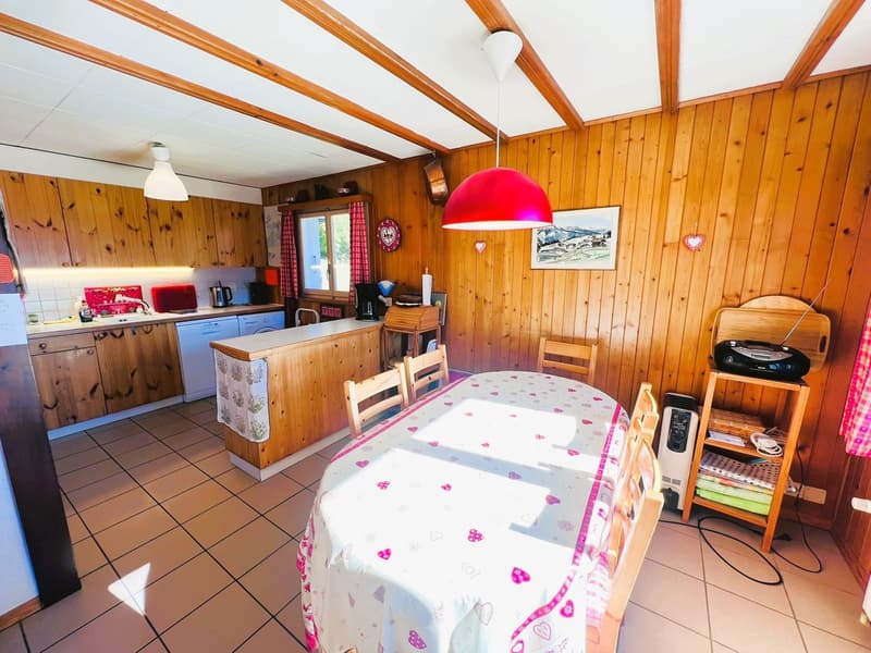 dining room, tiled floor, wooden ceiling beams, table with floral tablecloth, chairs, wooden kitchen counter with sink and appliances, wooden cabinets, kitchen island, picture frame, hanging lamp, shelf with radio and books