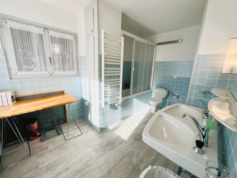 Bathroom with blue tiles, wooden desk, white sink, toilet, and glass windows.