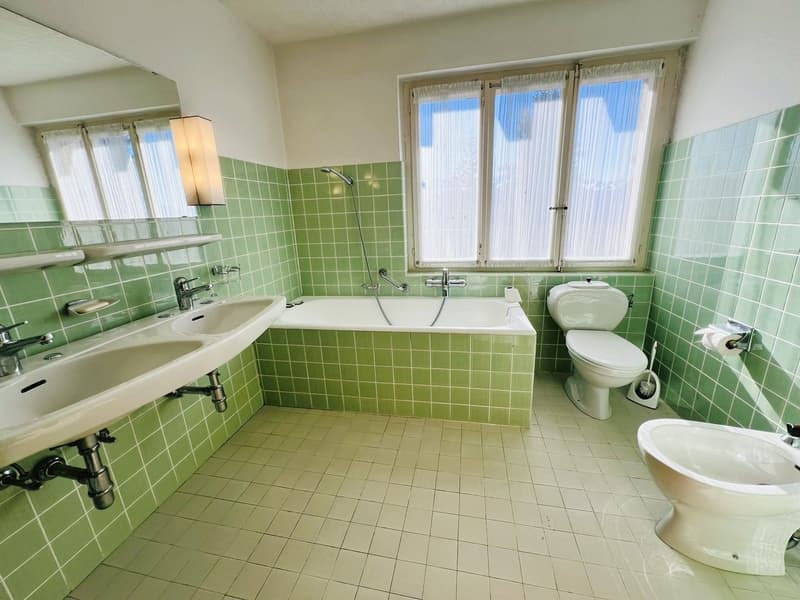 Green tiled walls, white tiled floor, double sink, bathtub, toilet, bidet, shower, window